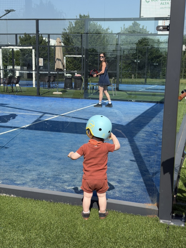 Toddler in a blue helmet watching his mom play padel tennis through the glass court wall on a sunny day in Italy