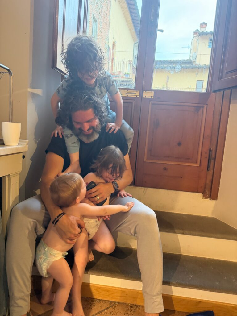 Dad laughing on apartment steps as four young boys in pajamas climb on him and hug him near a sunlit window overlooking Tuscan rooftops