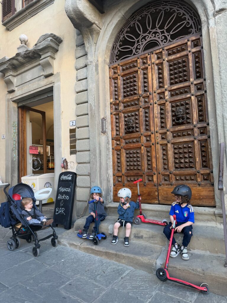 Three boys with scooters resting on ornate stone steps in Florence while toddler sits in stroller near a Bar Tabacchi sign