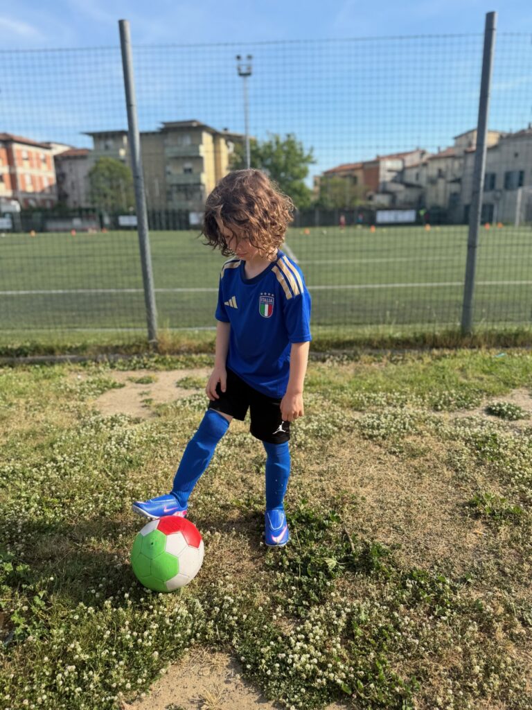 Young boy in a blue Italia national team jersey posing with a soccer ball on a grassy field in Italy
