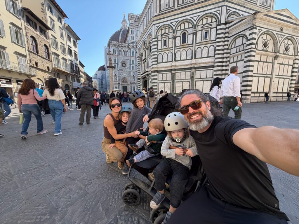 Family selfie with four boys in scooter helmets in front of the Florence Duomo and Baptistery in a bustling Italian piazza