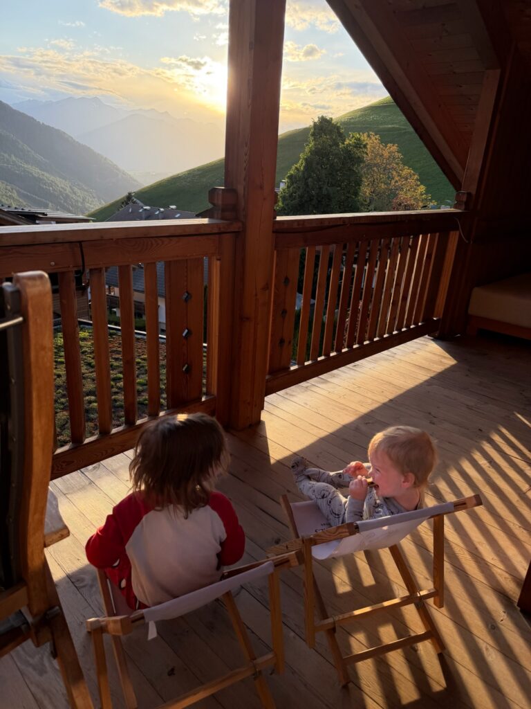 Two young boys eating snacks in wooden chairs on a Dolomites chalet balcony at sunset overlooking the Italian Alps