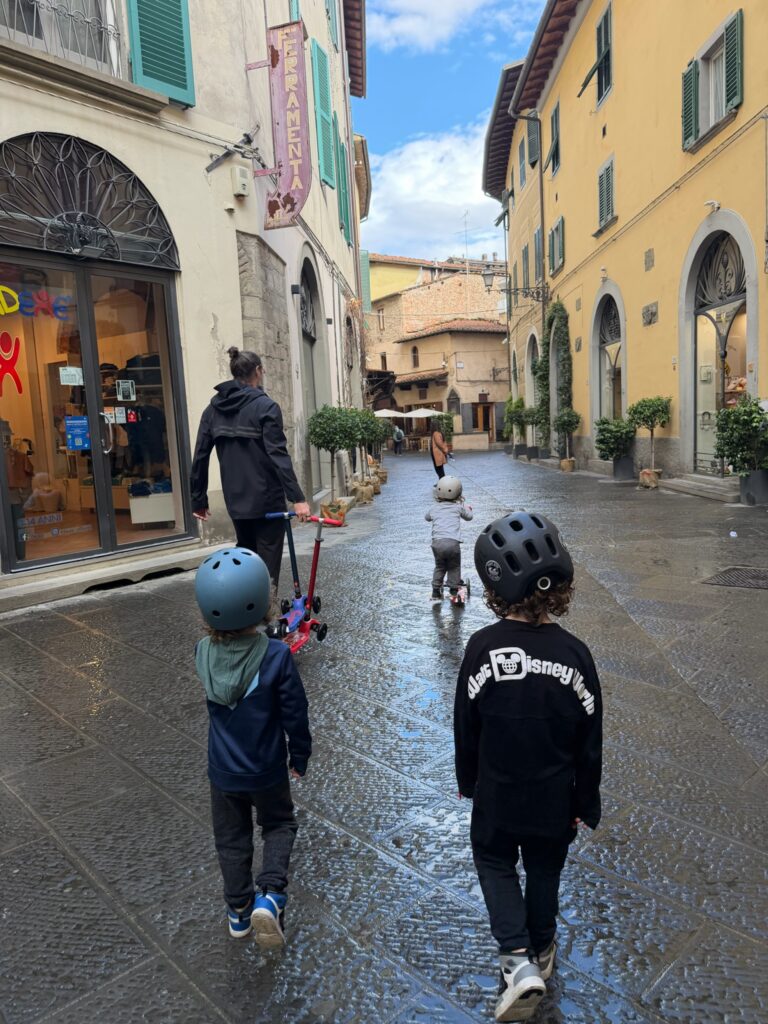 Dad and three boys in helmets riding scooters down a wet cobblestone street lined with pastel buildings in an Italian town