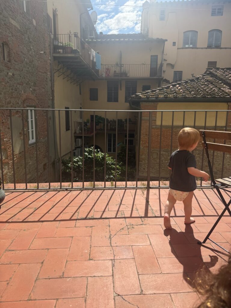Barefoot toddler exploring a sunlit terracotta balcony overlooking a traditional Tuscan courtyard with stone buildings and tiled roofs