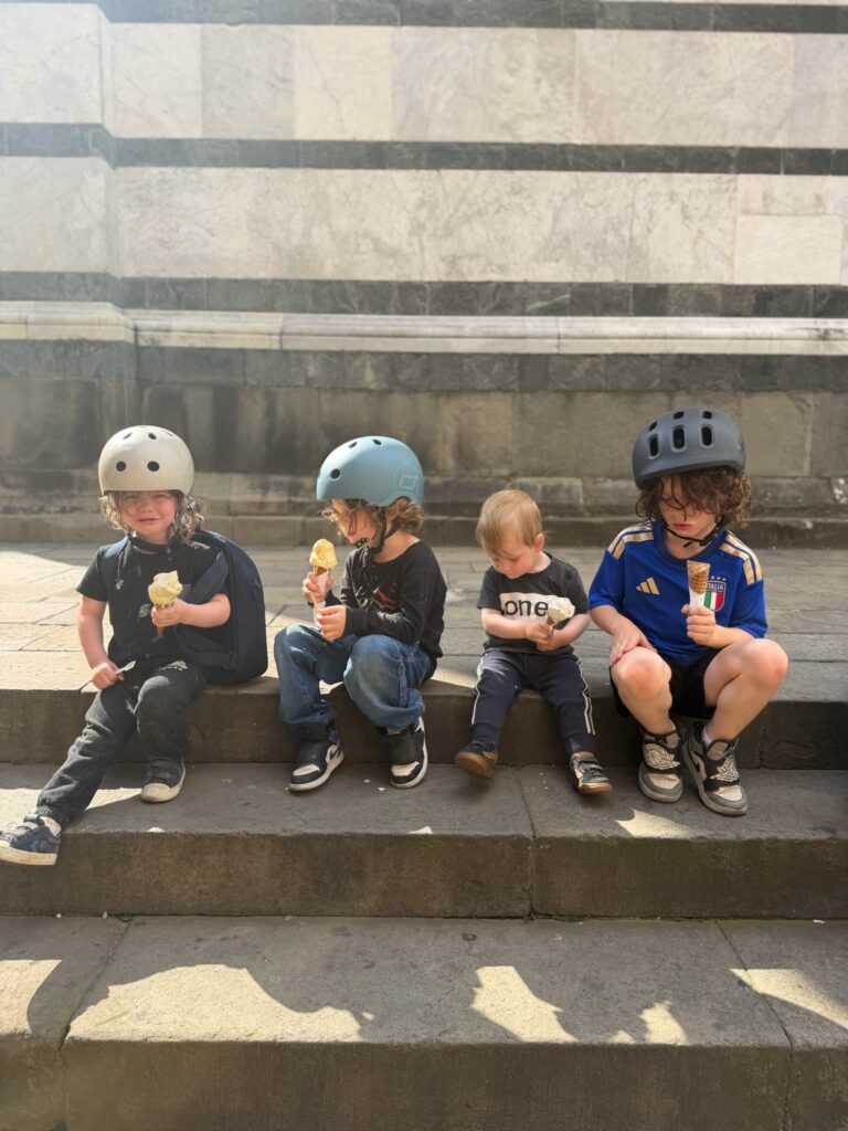 Four brothers in bike helmets sitting on marble cathedral steps eating gelato cones during a family day out in Italy