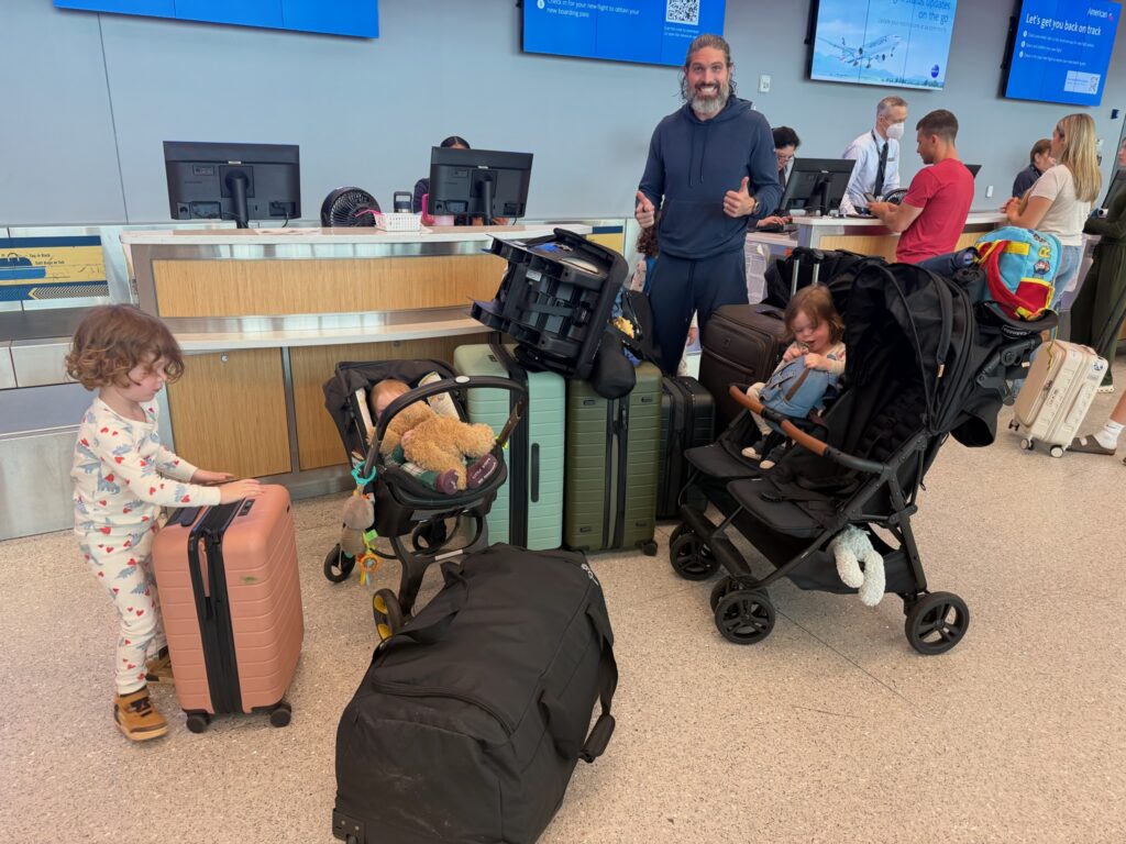 Dad gives thumbs up at American Airlines check-in counter surrounded by luggage, car seats, strollers, and four young boys ready for international family travel