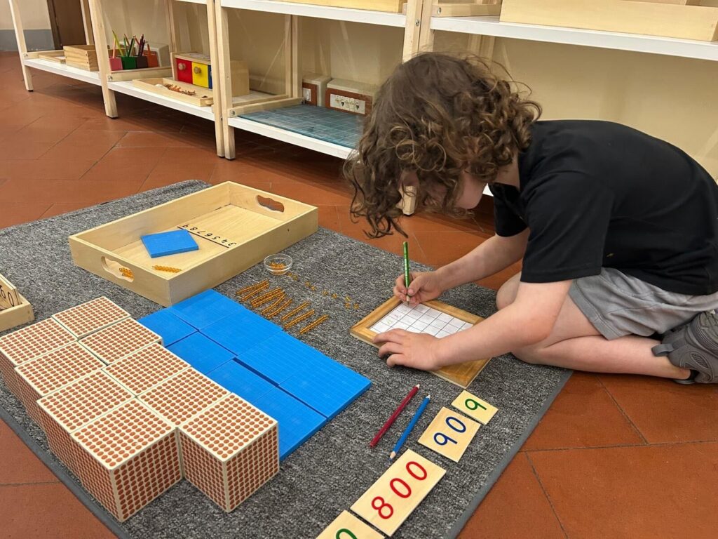 Curly-haired boy working with Montessori golden bead math materials on a classroom rug at Boundless Life world school in Italy
