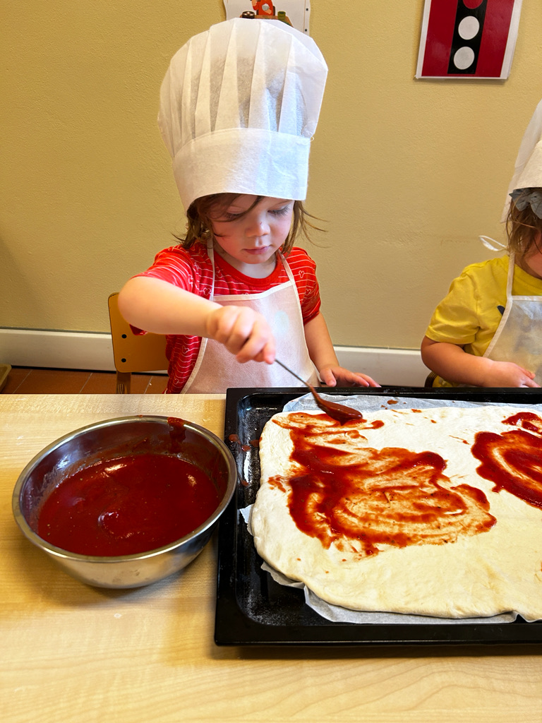 Young boy in a chef hat spreading tomato sauce on homemade pizza during a kids cooking class at Boundless Life in Italy